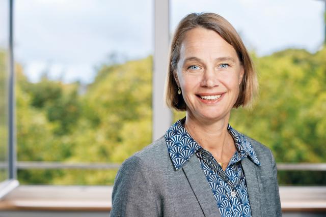 Eine Frau mit kurzen dunkelblonden Haaren lächelt in die Kamera und steht vor einem Fenster.