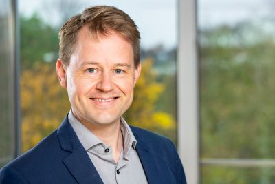 Ein Mann mit kurzen braunen Haaren lächelt in die Kamera und steht vor einem Fenster.