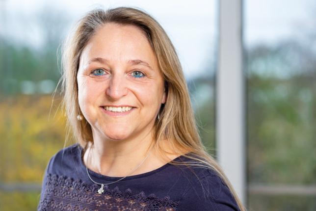 Eine Frau mit langen blonden Haaren und blauem Eyeliner lächelt in die Kamera und steht vor einem Fenster.