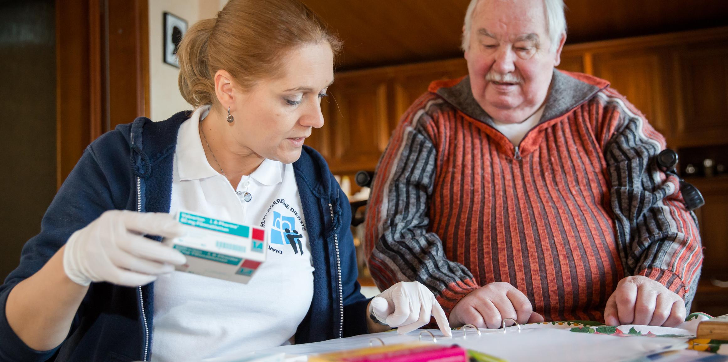 Eine Frau mit medizinischen Handschuhen hält eine Medikamentenpackung in der Hand und zeigt einem älteren Herren etwas in einem Ordner.