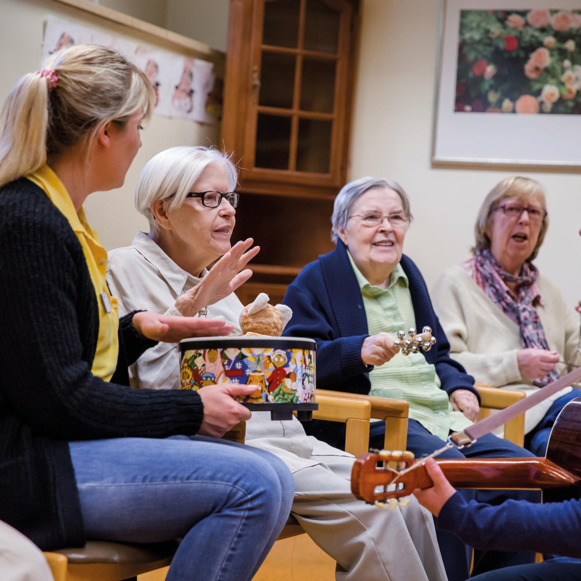 Eine Gruppe älterer Menschen sitzt gemeinsam mit zwei jüngeren Personen im Kreis und musiziert mit verschiedenen Instrumenten.
