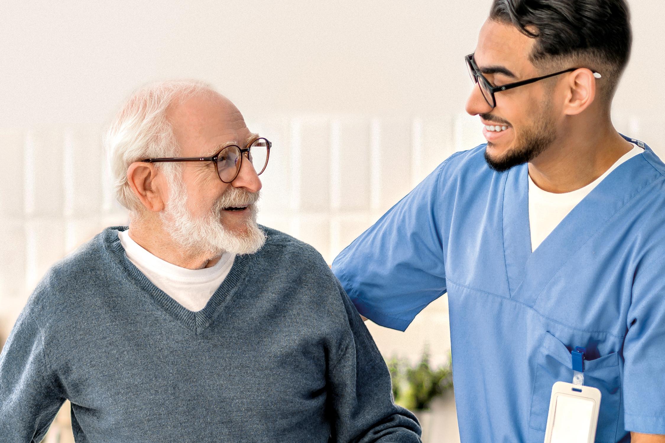 Smiling friendly in-home male nurse in uniform supporting an old man with a walking stick