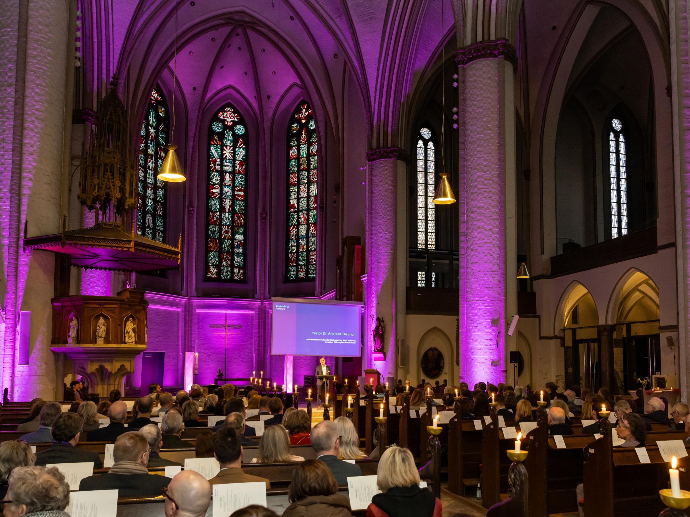 Innenaufnahme einer großen Kirche während einer Veranstaltung. Viele Menschen sitzen in den Bänken, Kerzen stehen an den Seiten. Der Chorraum ist in violettem Licht ausgeleuchtet, vorne ist eine Person am Redepult zu sehen. Über dem Altarraum befinden sich große bunte Kirchenfenster und gotische Gewölbebögen.