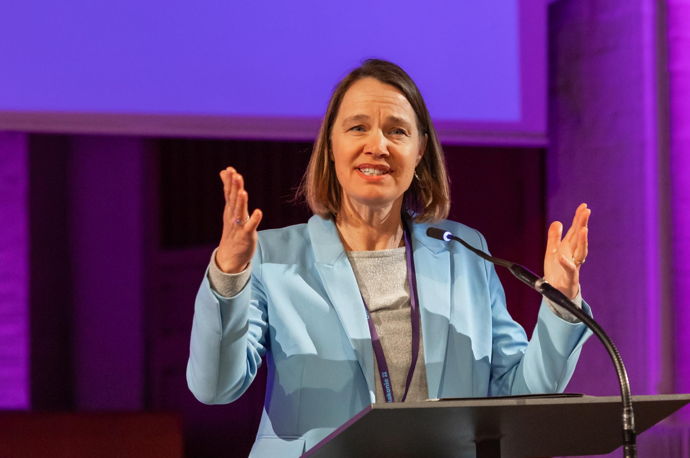 Eine Person steht an einem Redepult und spricht mit ausdrucksvollen, erhobenen Händen. Sie trägt ein hellblaues Sakko über einem grauen Oberteil. Das Umfeld ist in violettes Licht getaucht, im Hintergrund sind architektonische Elemente eines großen Raumes zu erkennen.