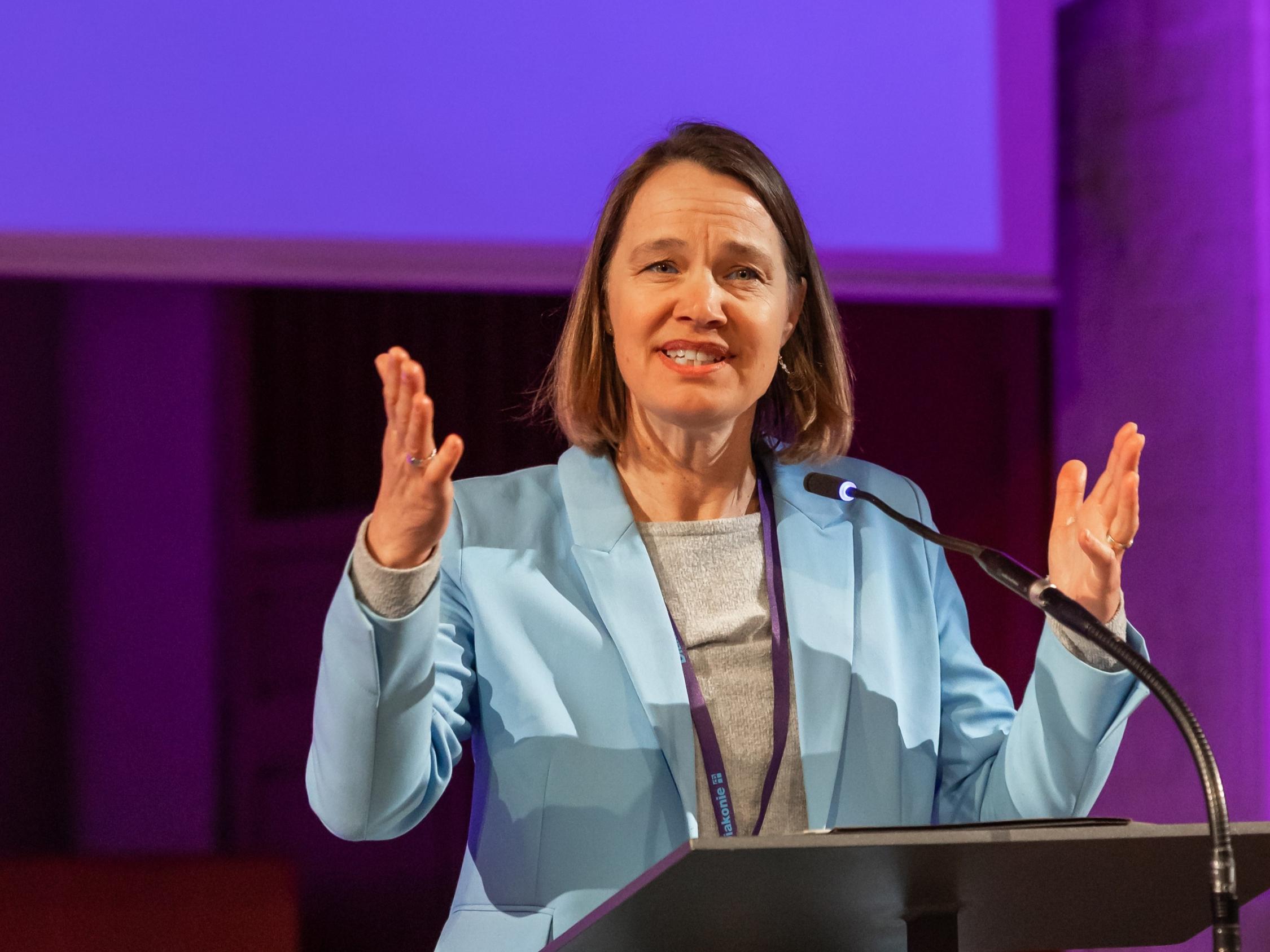 Eine Person steht an einem Redepult und spricht mit ausdrucksvollen, erhobenen Händen. Sie trägt ein hellblaues Sakko über einem grauen Oberteil. Das Umfeld ist in violettes Licht getaucht, im Hintergrund sind architektonische Elemente eines großen Raumes zu erkennen.