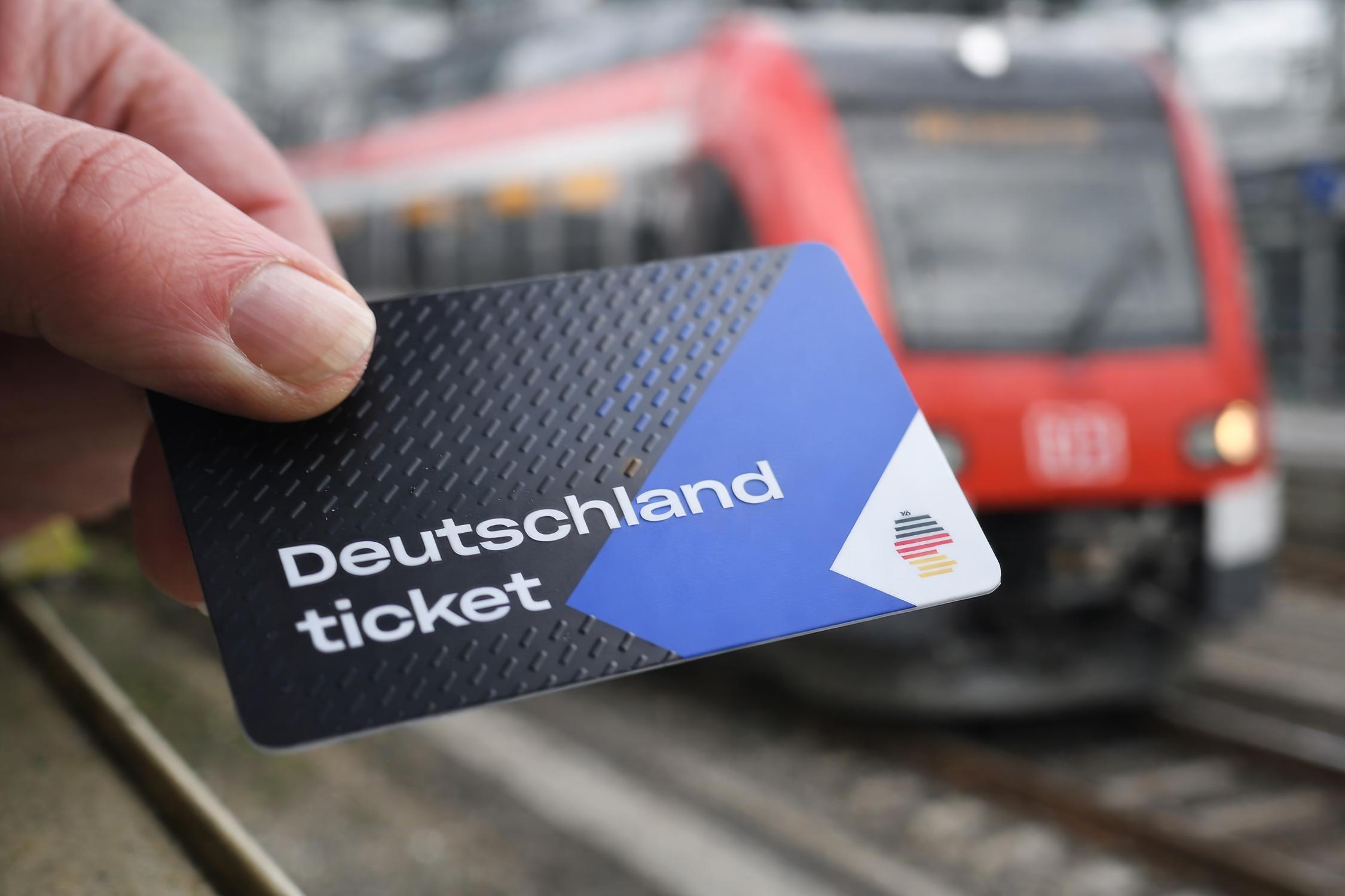 Eine blau-weiß-schwarze Plastikkarte in einer menschlichen Hand. im Hintergrund ist eine Hamburger S-Bahn zu sehen.