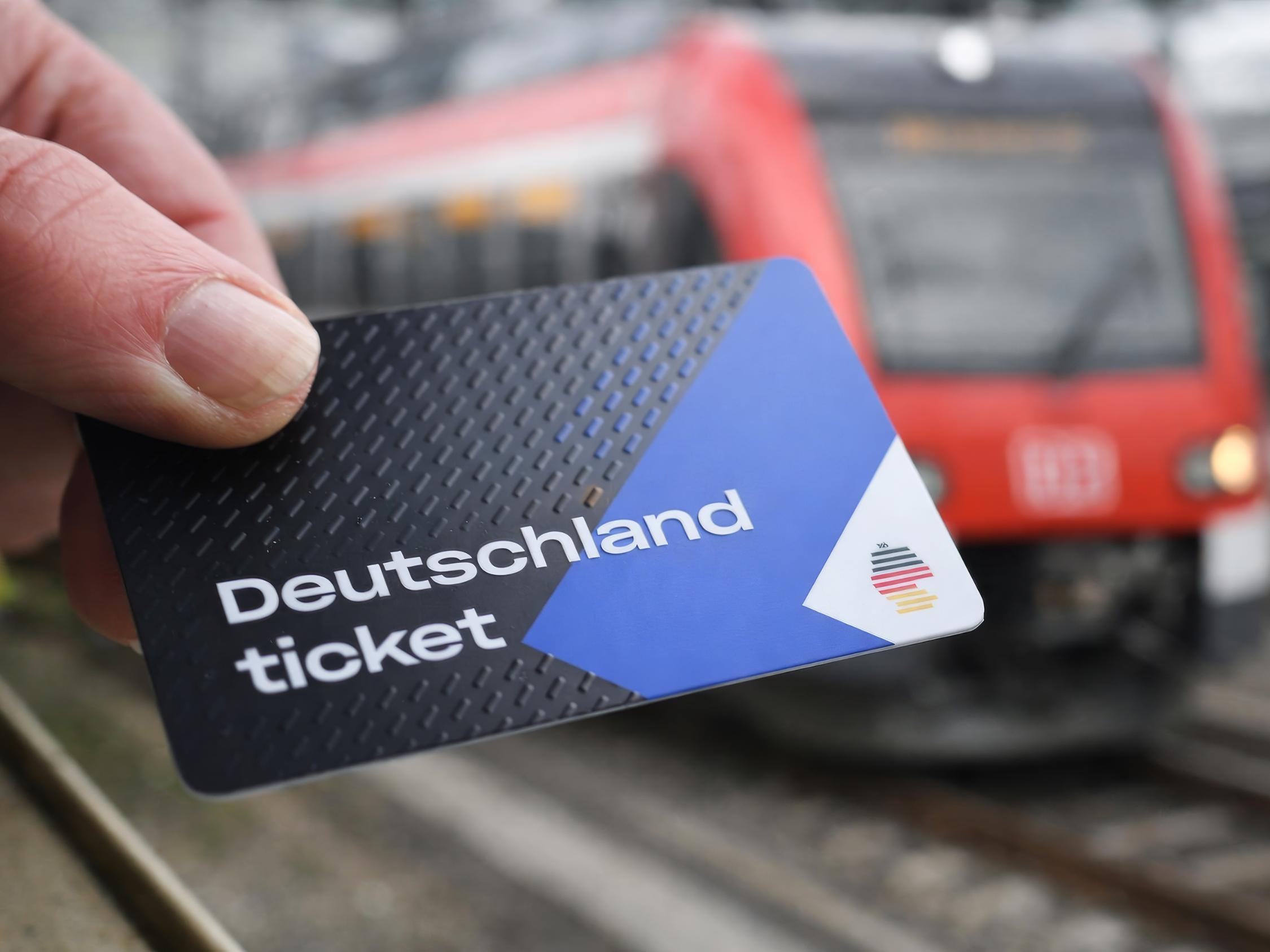 Eine blau-weiß-schwarze Plastikkarte in einer menschlichen Hand. im Hintergrund ist eine Hamburger S-Bahn zu sehen.