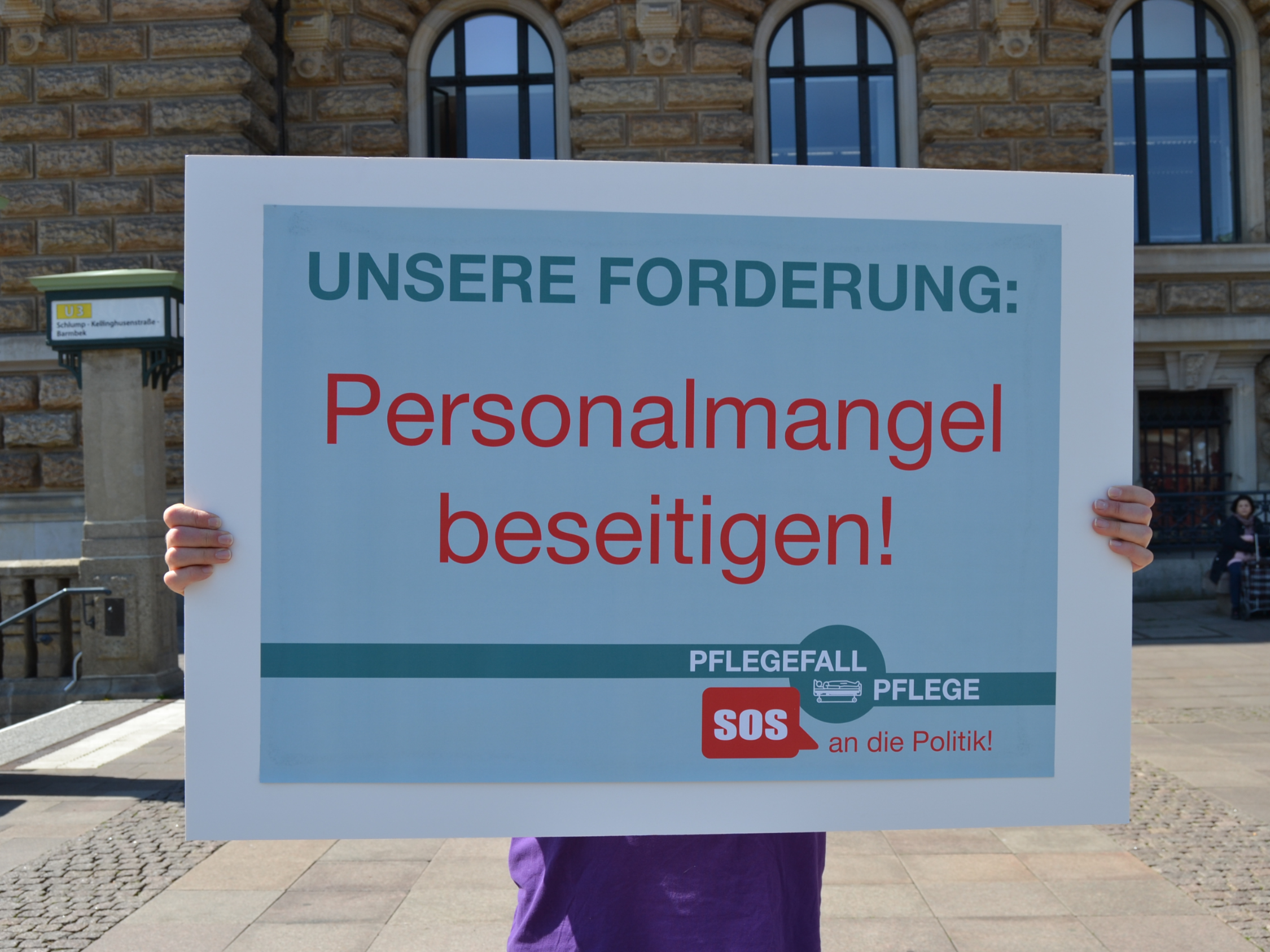 Eine Person hält ein Schild hoch, auf dem die Beseitigung des Personalmangels in der Pflege gefordert wird.