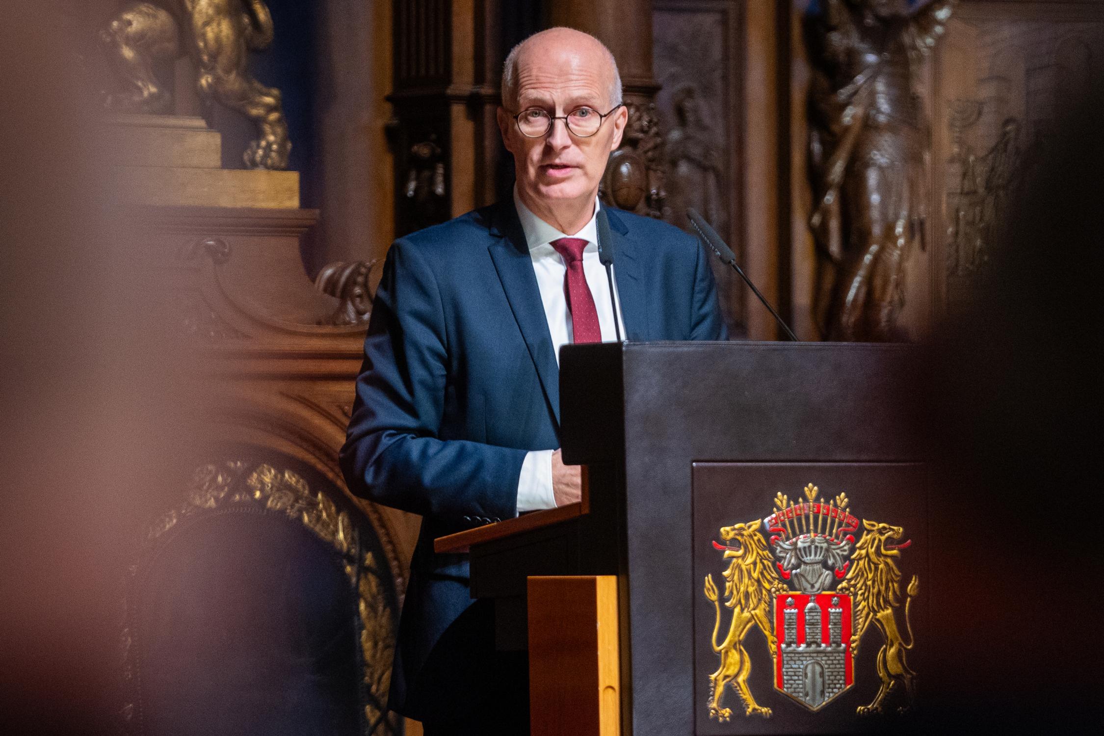Ein Mann mit weißen Haaren und einer Brille steht an einem Pult und spricht in ein Mikrofon.