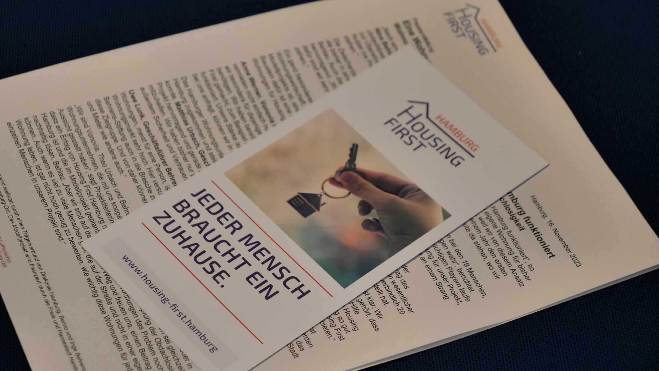 Auf einem Handout zu 'Housing First Hamburg' liegt eine Broschüre mit dem Titel 'Jeder Mensch braucht ein Zuhause