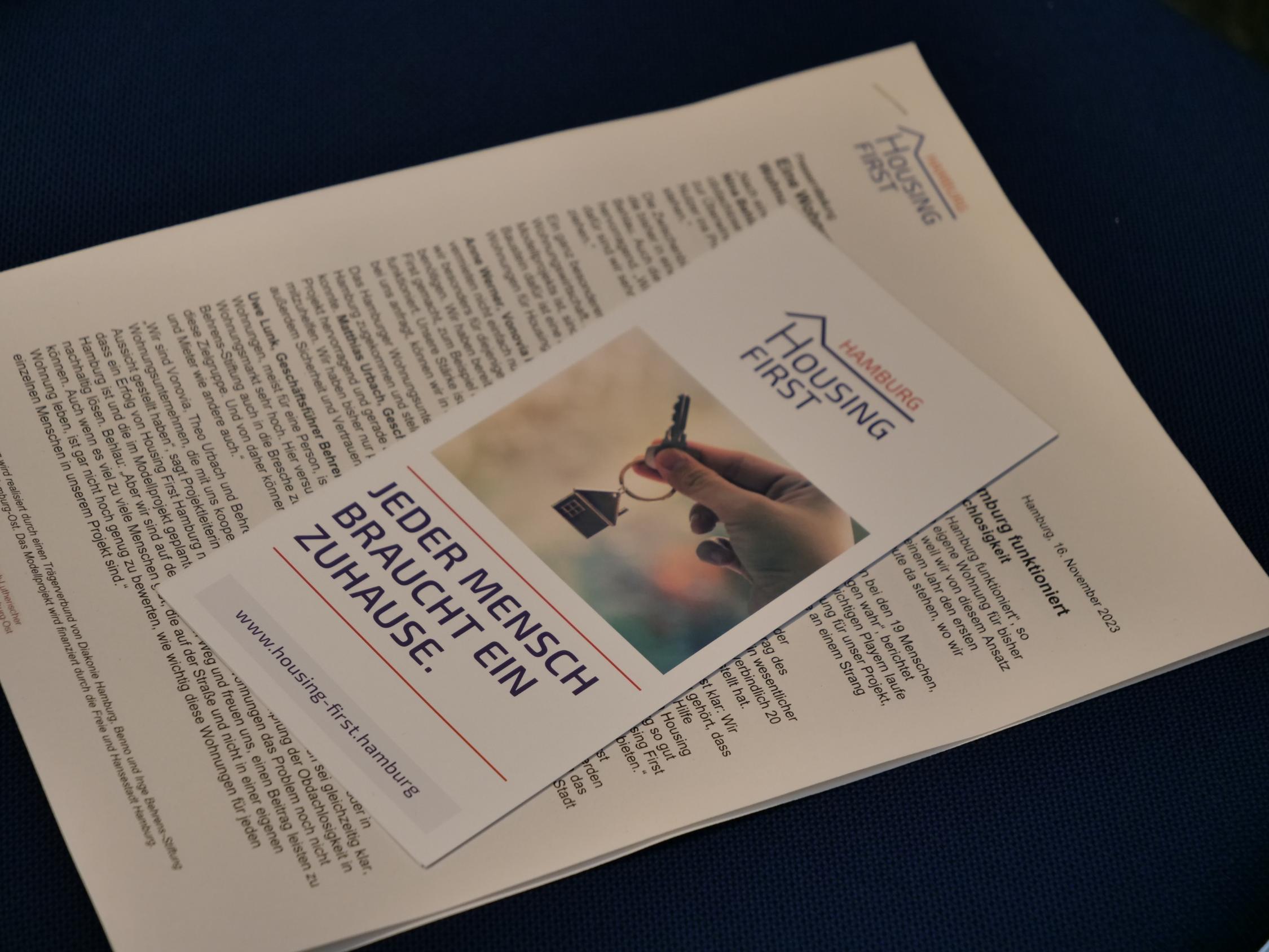 Auf einem Handout zu 'Housing First Hamburg' liegt eine Broschüre mit dem Titel 'Jeder Mensch braucht ein Zuhause