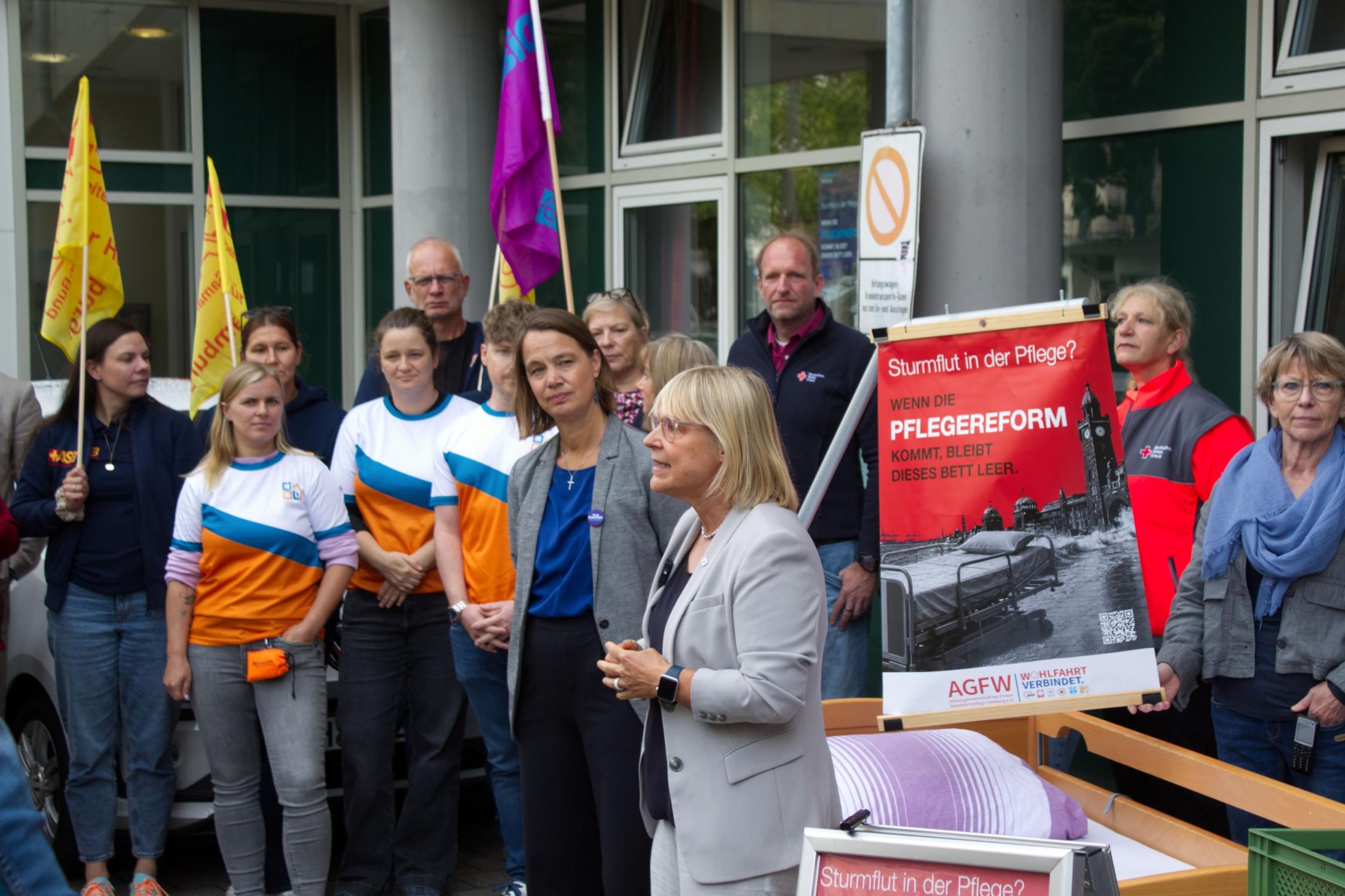 Das Foto zeigt eine öffentlichkeitswirksame Aktion vor dem Eingang eines größeren Gebäudes. Im Mittelpunkt steht ein Pflegebett, das im Vordergrund aufgestellt wurde und als zentrales Symbol der Veranstaltung dient. Um das Bett herum haben sich etwa ein Dutzend Personen versammelt. Einige tragen Arbeitskleidung oder Westen von Organisationen, andere sind in ziviler Kleidung erschienen. Mehrere Teilnehmende halten Fahnen in Gelb und Violett.
