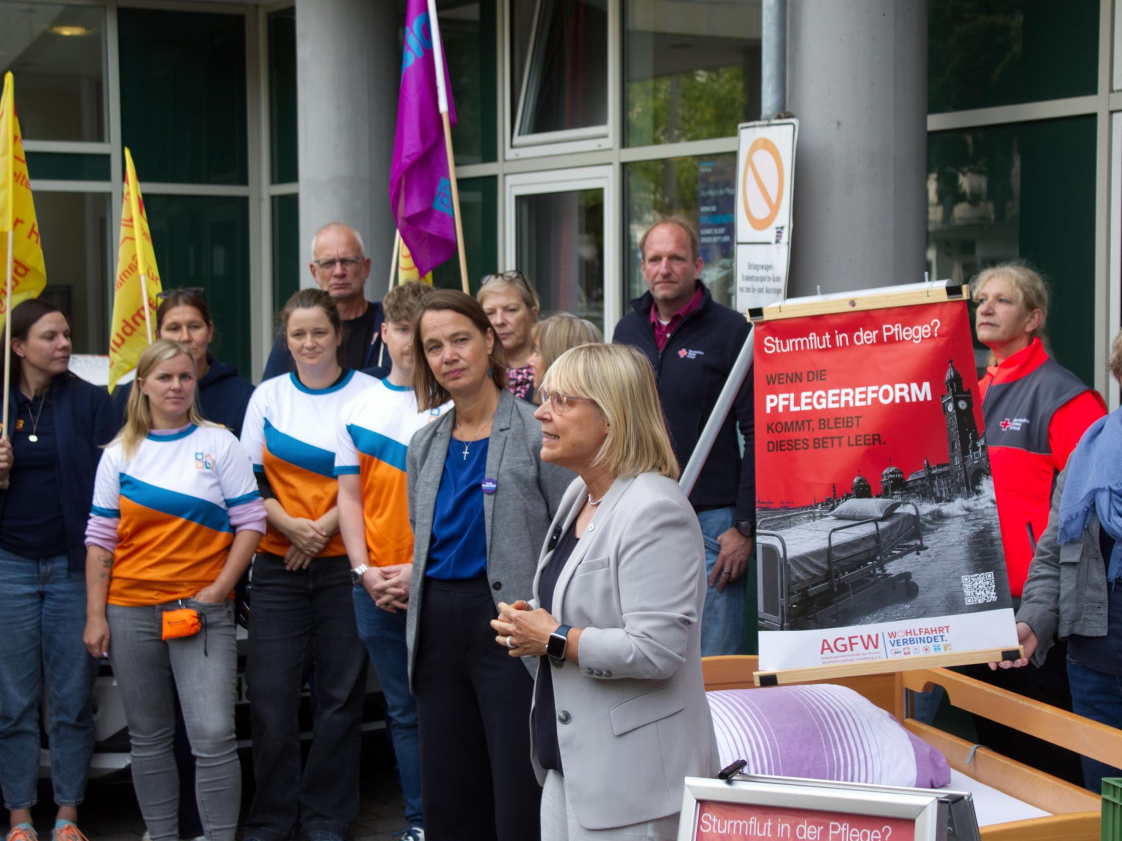 Das Foto zeigt eine öffentlichkeitswirksame Aktion vor dem Eingang eines größeren Gebäudes. Im Mittelpunkt steht ein Pflegebett, das im Vordergrund aufgestellt wurde und als zentrales Symbol der Veranstaltung dient. Um das Bett herum haben sich etwa ein Dutzend Personen versammelt. Einige tragen Arbeitskleidung oder Westen von Organisationen, andere sind in ziviler Kleidung erschienen. Mehrere Teilnehmende halten Fahnen in Gelb und Violett.