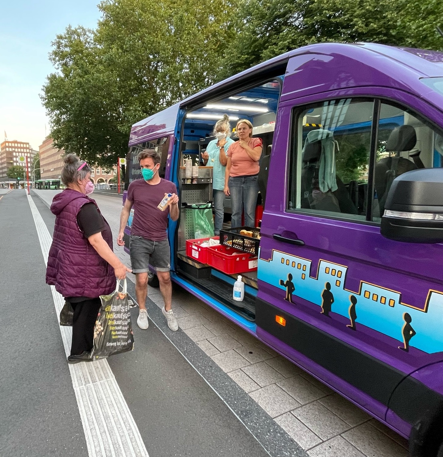 Ein Mann mit Maske steht vor dem lila Mitternachtsbus und hält einer Frau mit Tüten Sonnencreme entgegen.