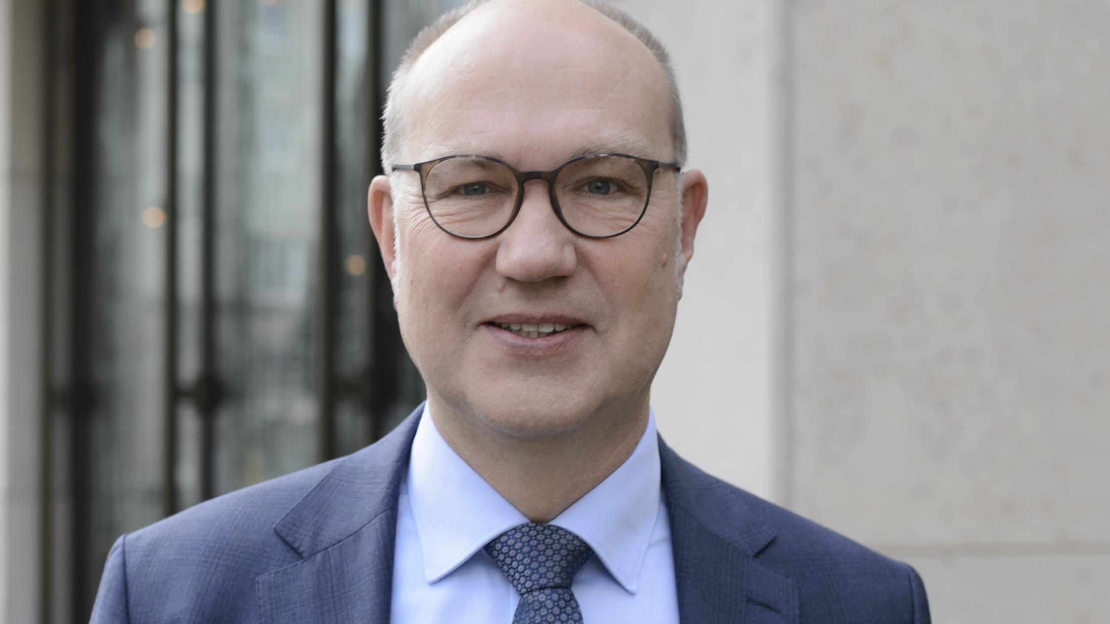 Der Oberkörper eines Mannes im Anzug ist zu sehen. Er schaut direkt in die Kamera und trägt eine Brille.