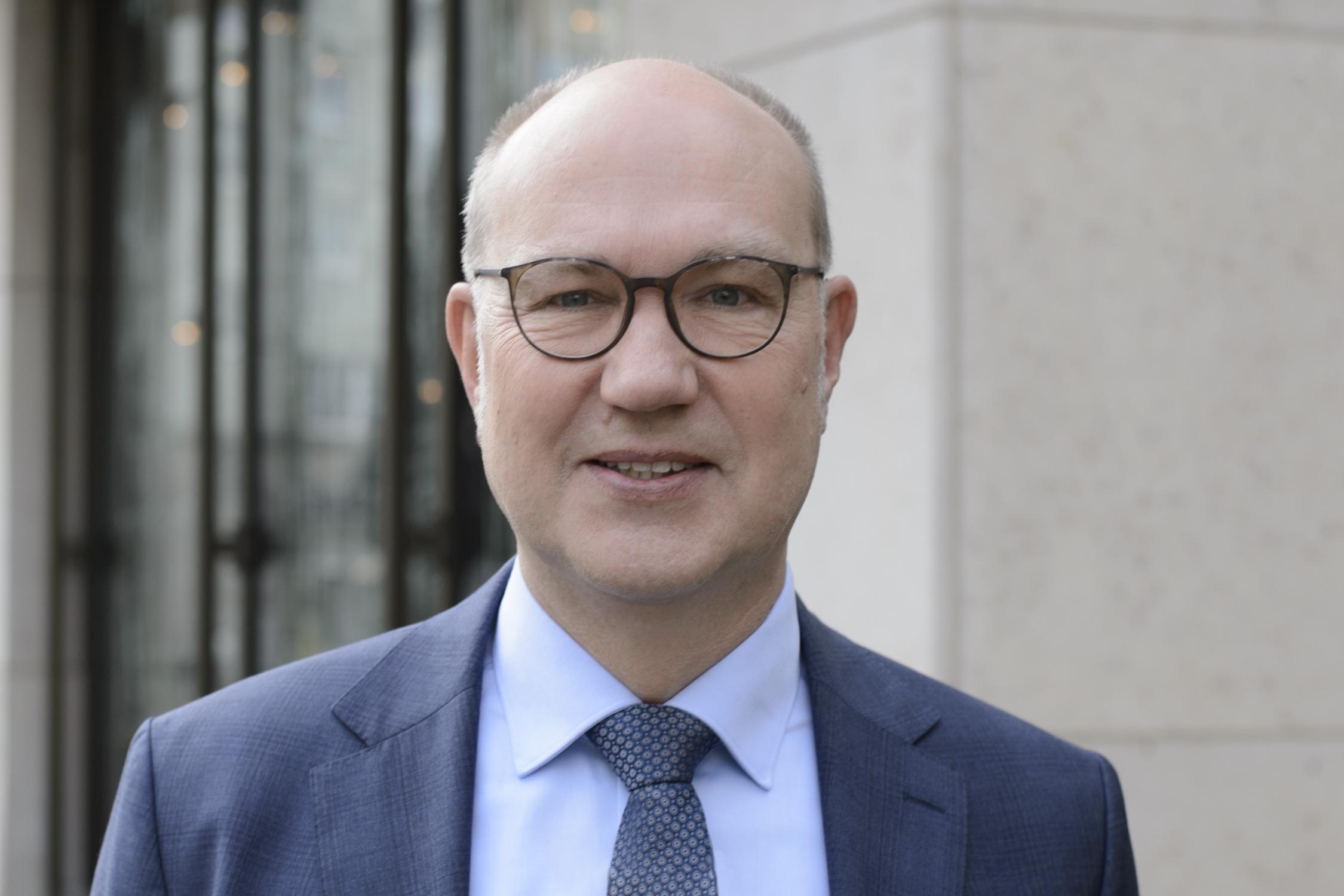 Der Oberkörper eines Mannes im Anzug ist zu sehen. Er schaut direkt in die Kamera und trägt eine Brille.