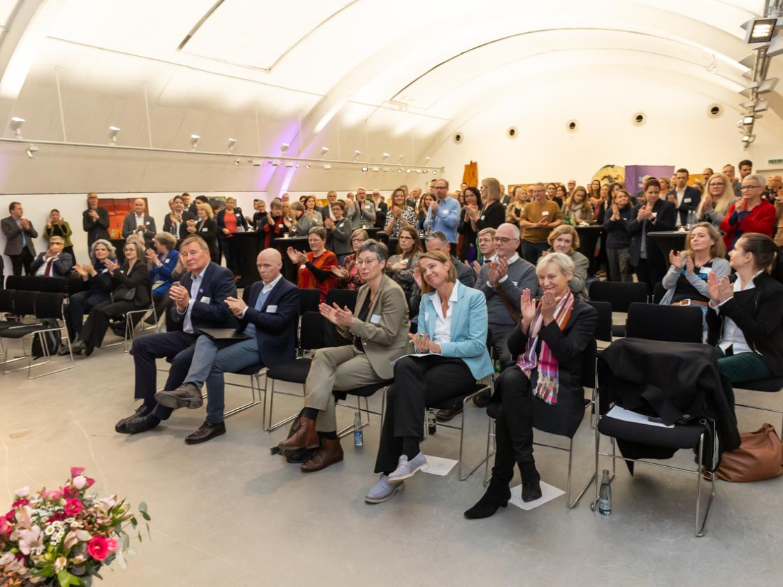 In einem Saal mit gewölbter Decke sitzen viele Menschen in Stuhlreihen und applaudieren.