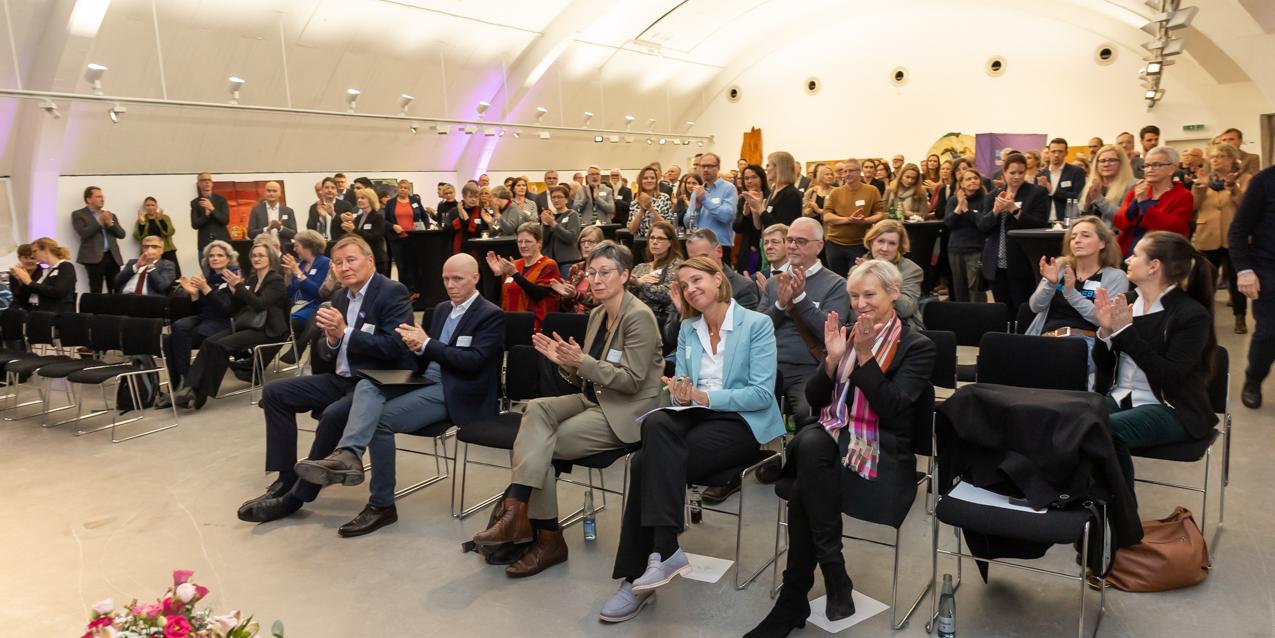 In einem Saal mit gewölbter Decke sitzen viele Menschen in Stuhlreihen und applaudieren.