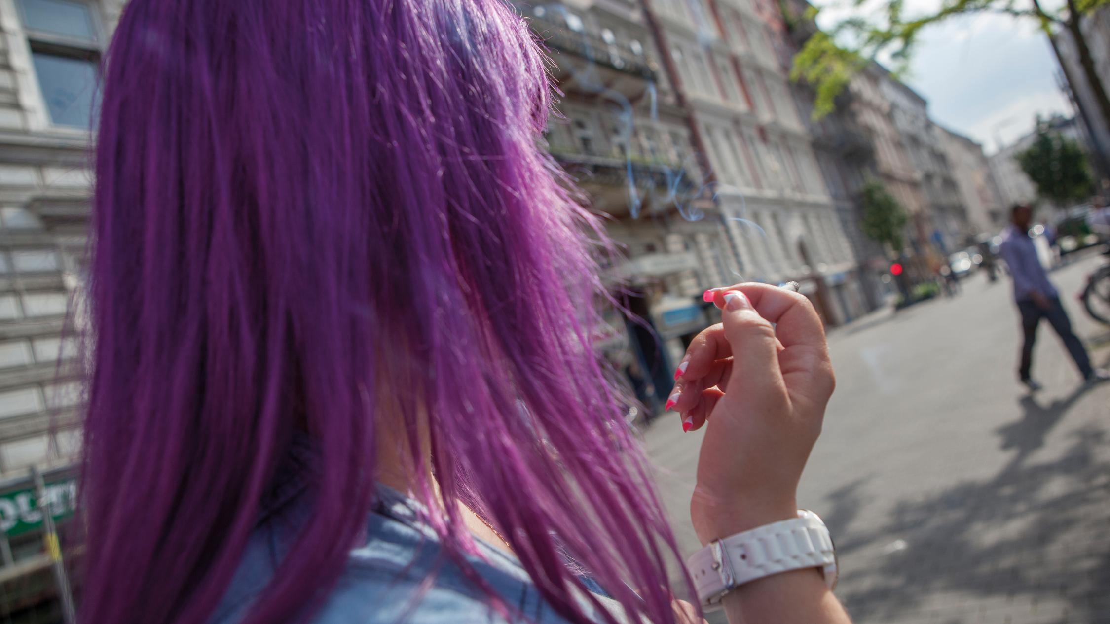 Man sieht den Hinterkopf einer rauchenden Frau mit violetten Haaren, die auf die Straße blickt.