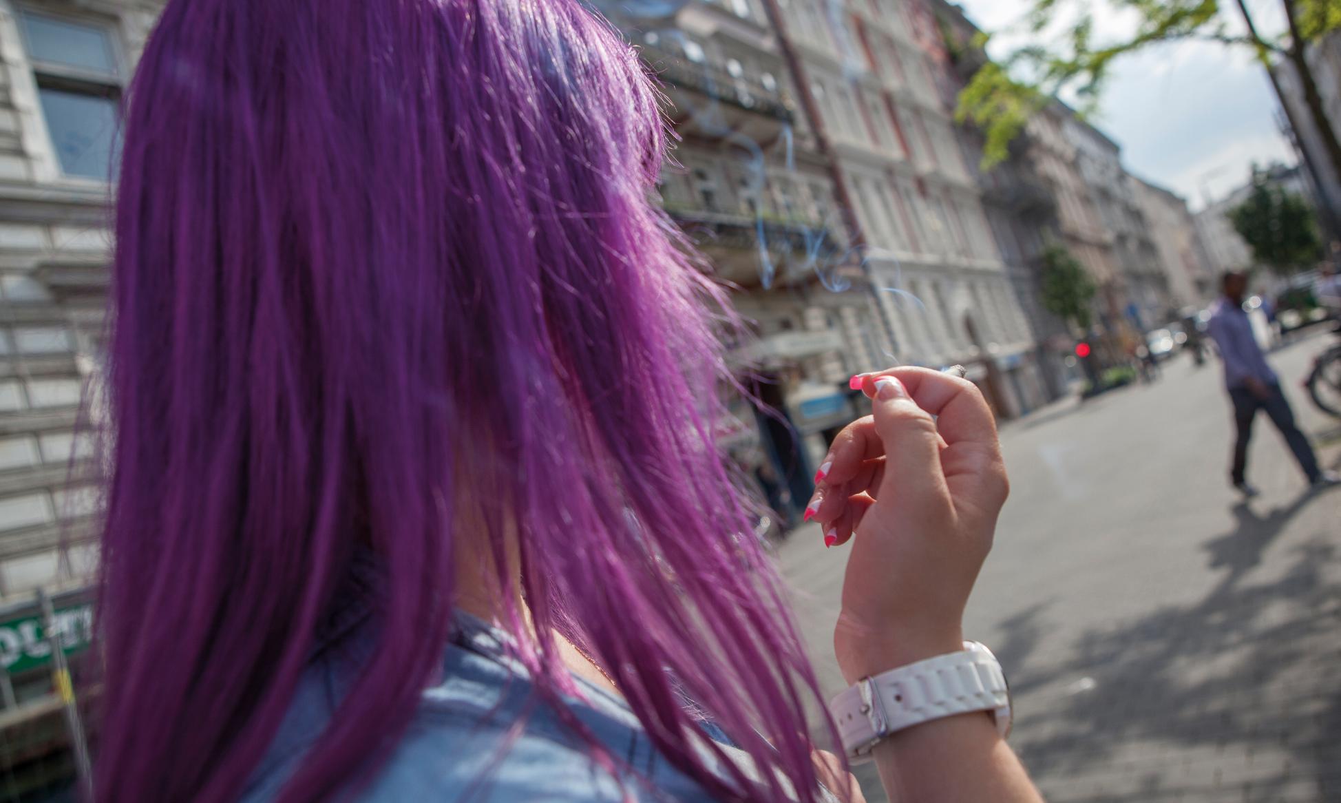 Man sieht den Hinterkopf einer rauchenden Frau mit violetten Haaren, die auf die Straße blickt.