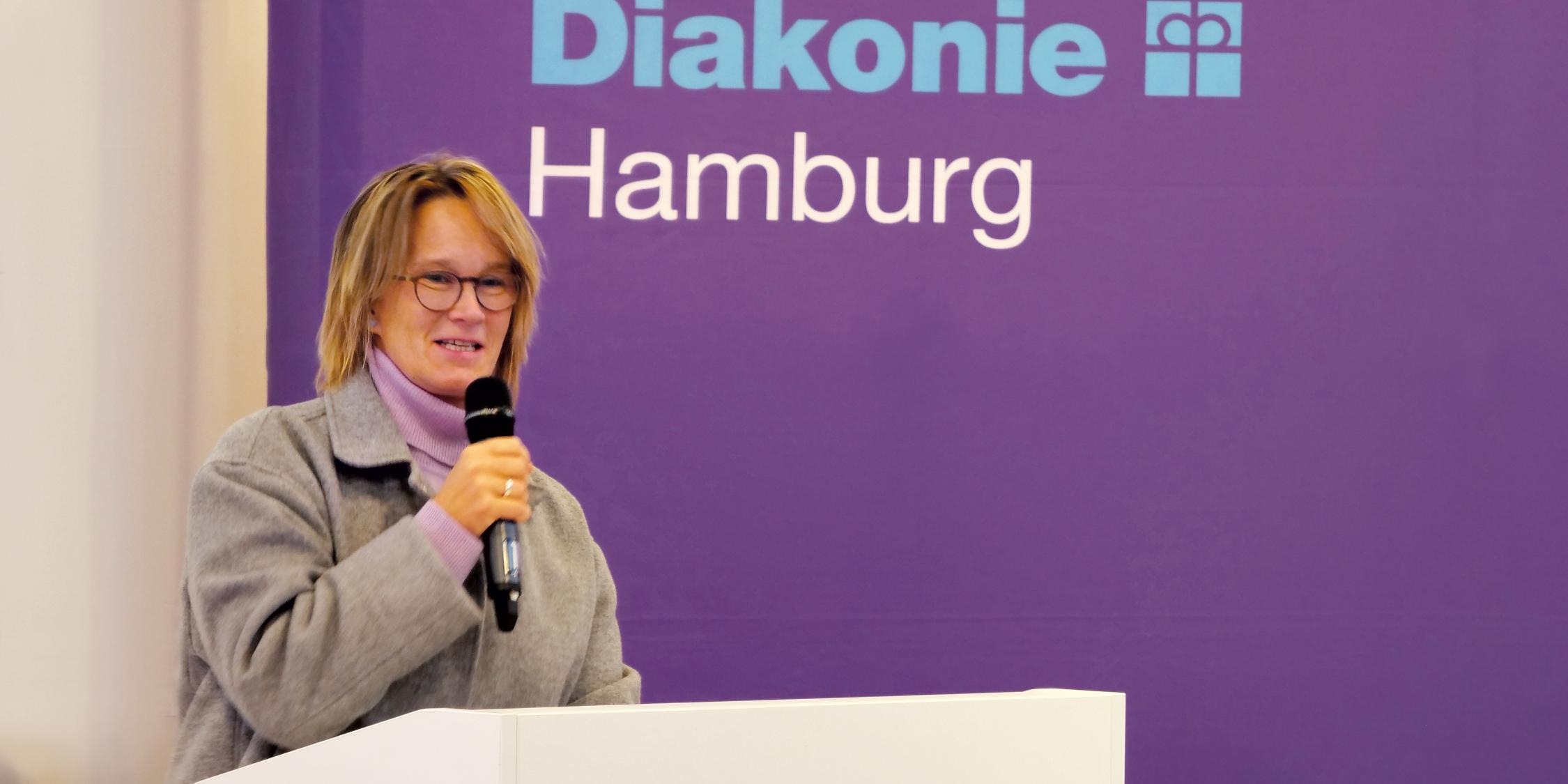 Eine blonde Frau steht vor einem Plakat der Diakonie an einem Pult und spricht in ein Mikrofon in ihrer Hand.