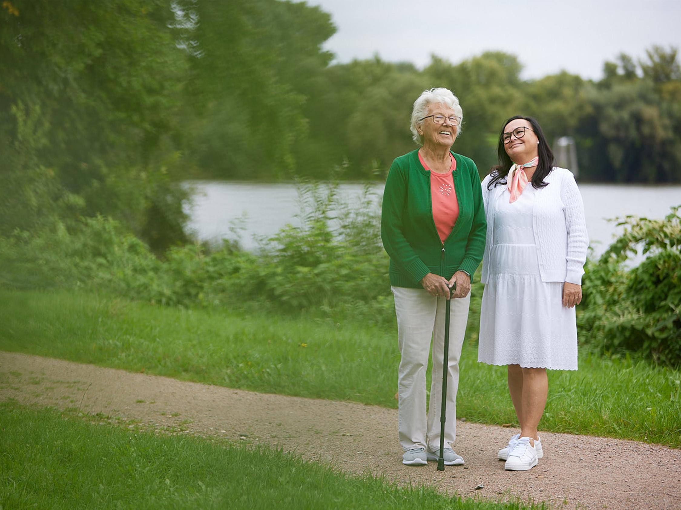 Eine ältere Frau mit Gehstock und eine jüngere Frau im Kleid stehen lächelnd nebeneinander an einem See.