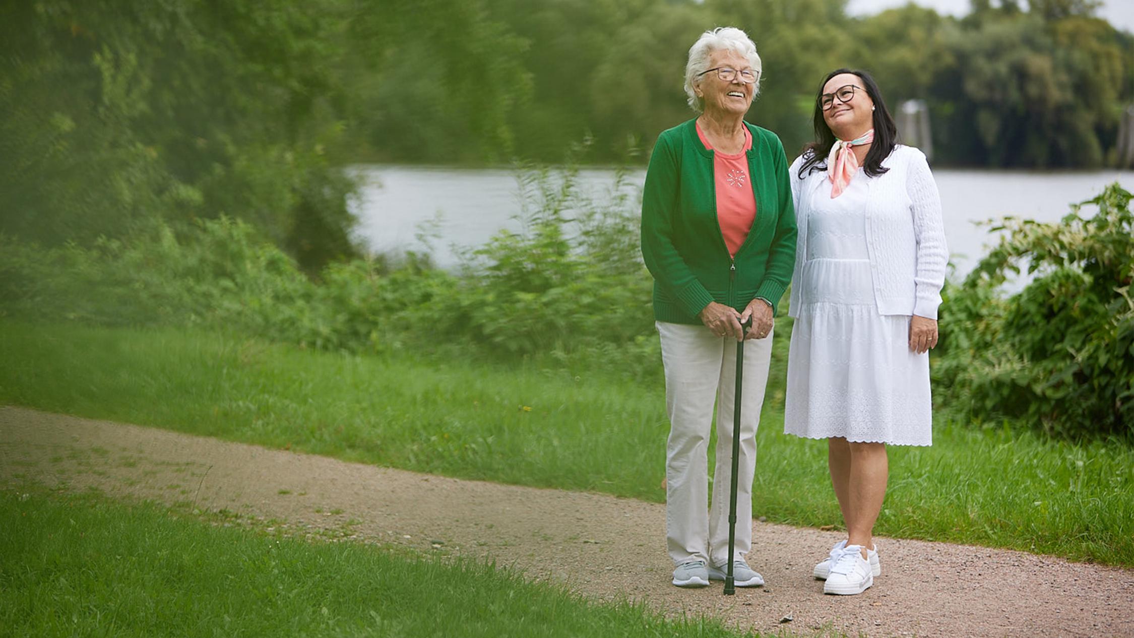 Eine ältere Frau mit Gehstock und eine jüngere Frau im Kleid stehen lächelnd nebeneinander an einem See.