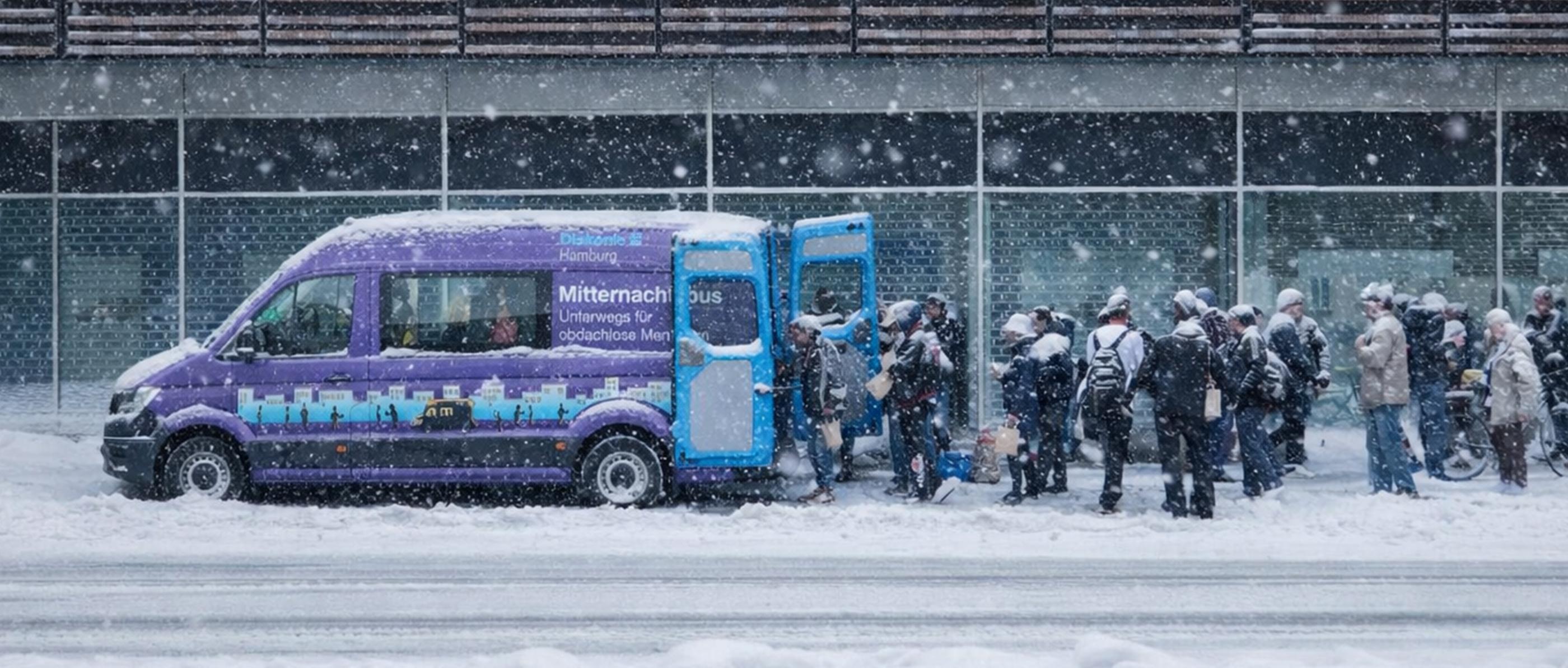 Menschen bilden eine Schlange hinter dem verschneiten lila Bus der Diakonie.