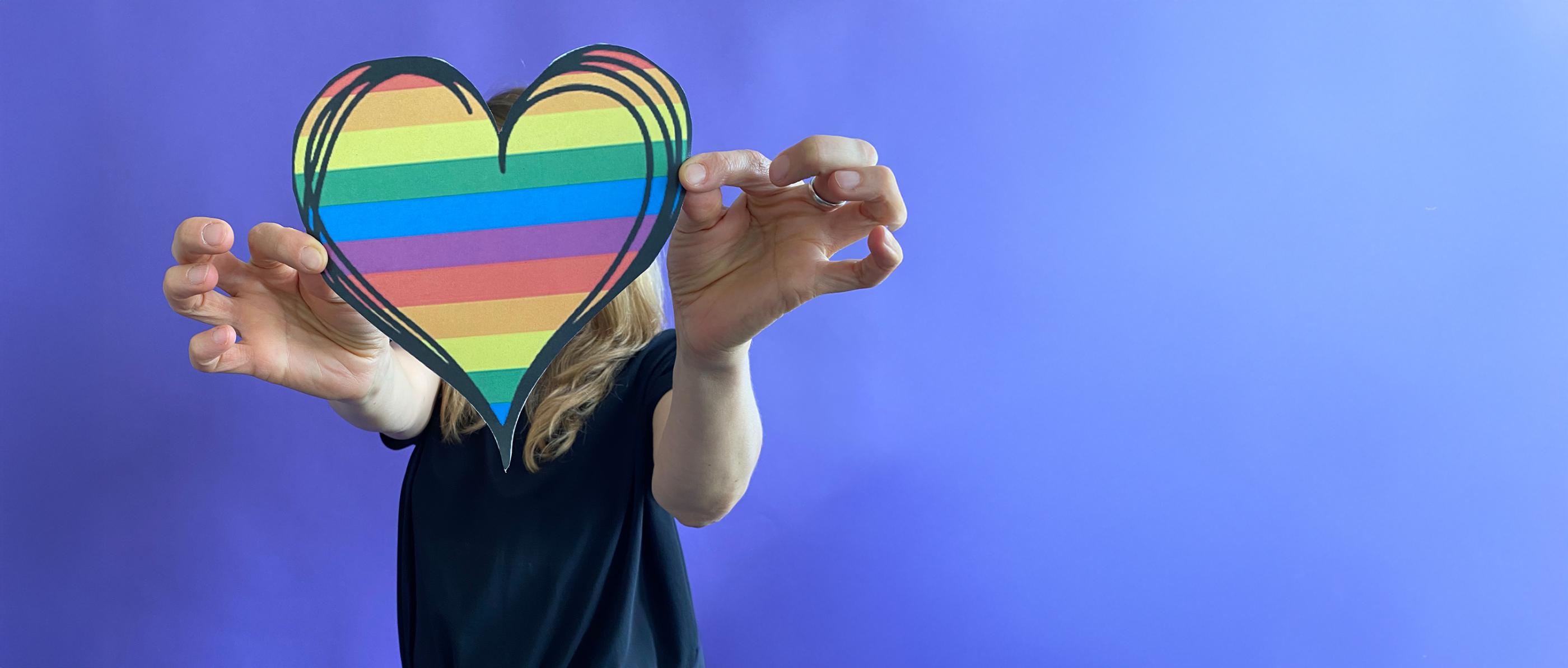 Eine Frau hält ein Herz in Regenbogenfarben in beiden Händen vor ihrem Kopf