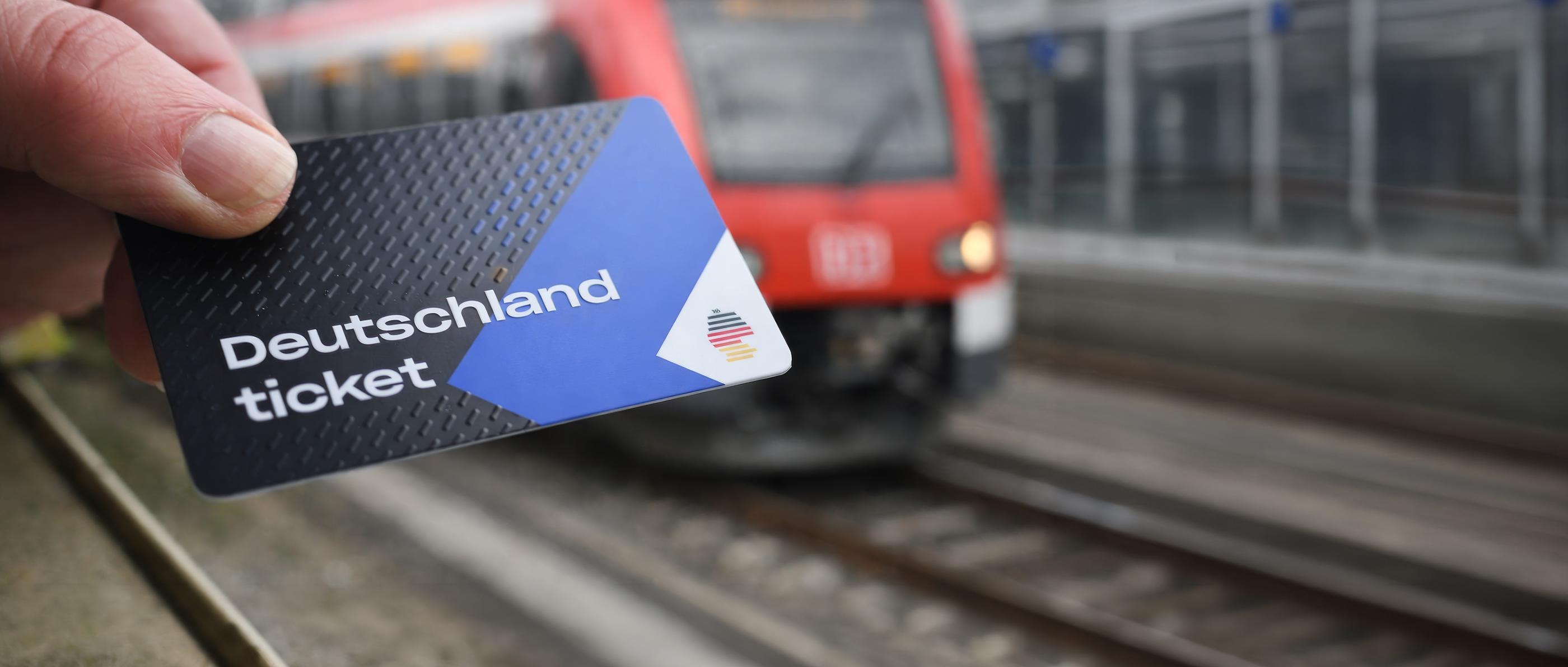 Eine blau-weiß-schwarze Plastikkarte in einer menschlichen Hand. im Hintergrund ist eine Hamburger S-Bahn zu sehen.