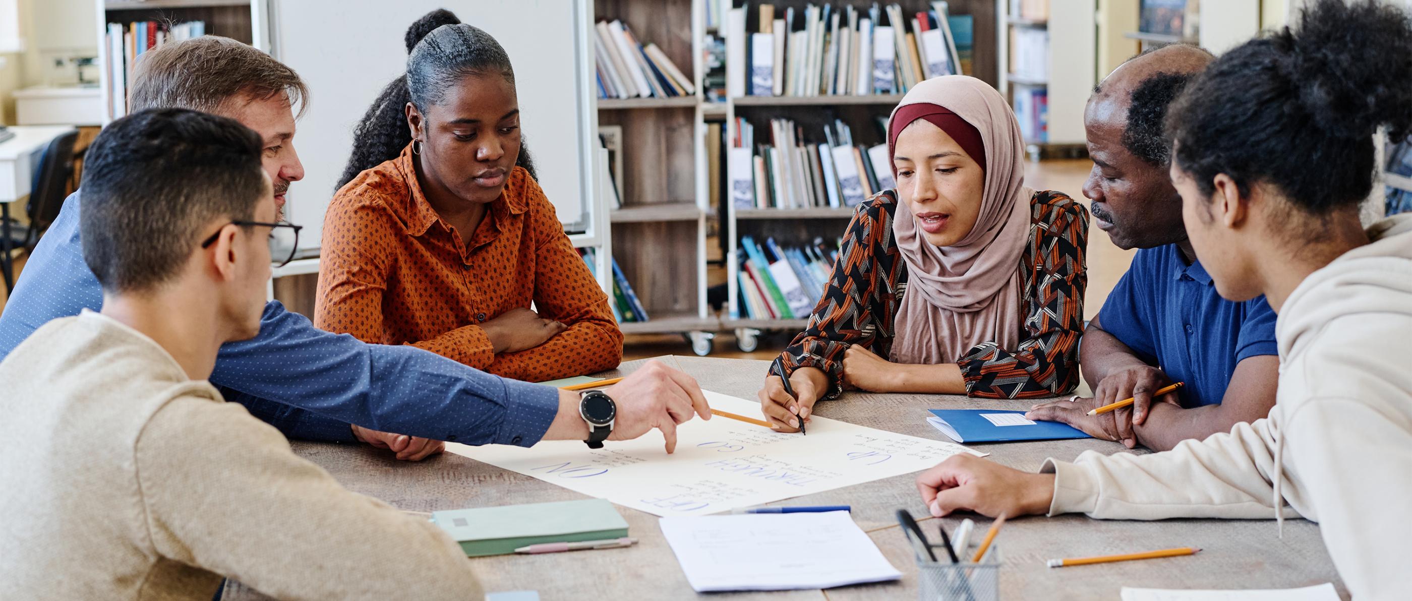 Sechs Menschen mutmaßlich unterschiedlicher Herkunft sitzen an einem Tisch.