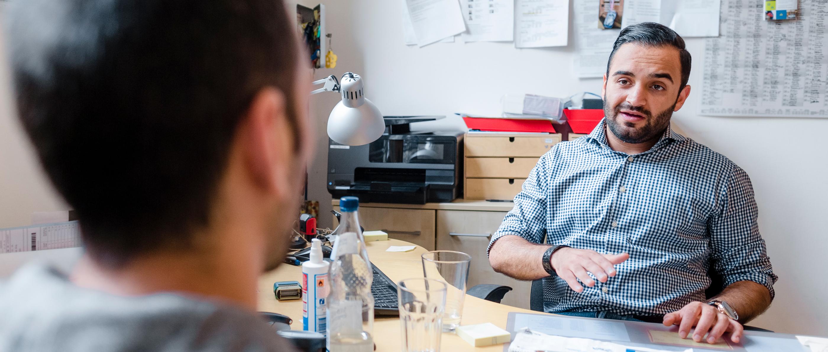 Ein Mann sitzt an einem Schreibtisch in einem Büro und spricht mit einem anderen Mann, der ihm gegenüber sitzt.