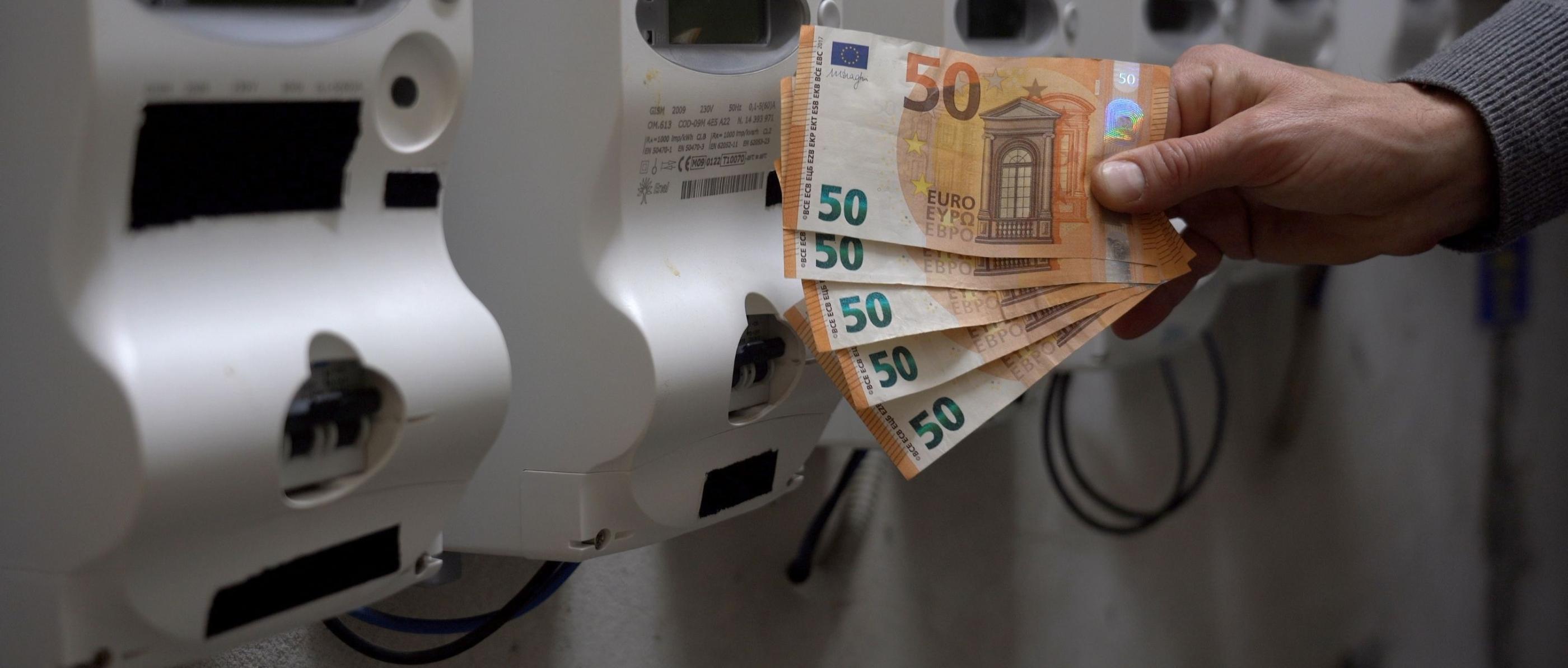 Eine Hand hält mehrere fünfzig Euro Scheine. Im Hintergrund sind Stromzähler zu sehen.