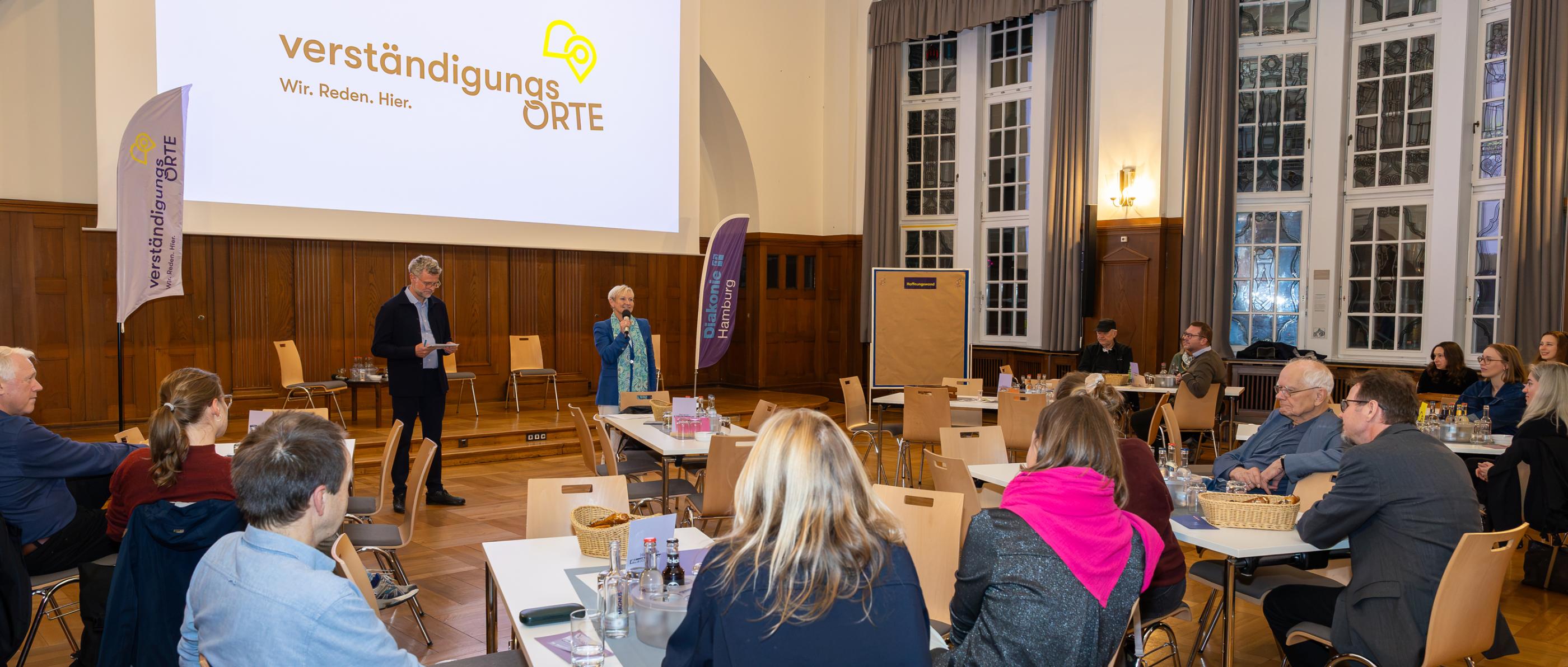 Menschen sitzen in einem Saal. Im Hintergrund steht eine Frau mit einem Mikrophon in der Hand. Neben ihr steht ein Mann.