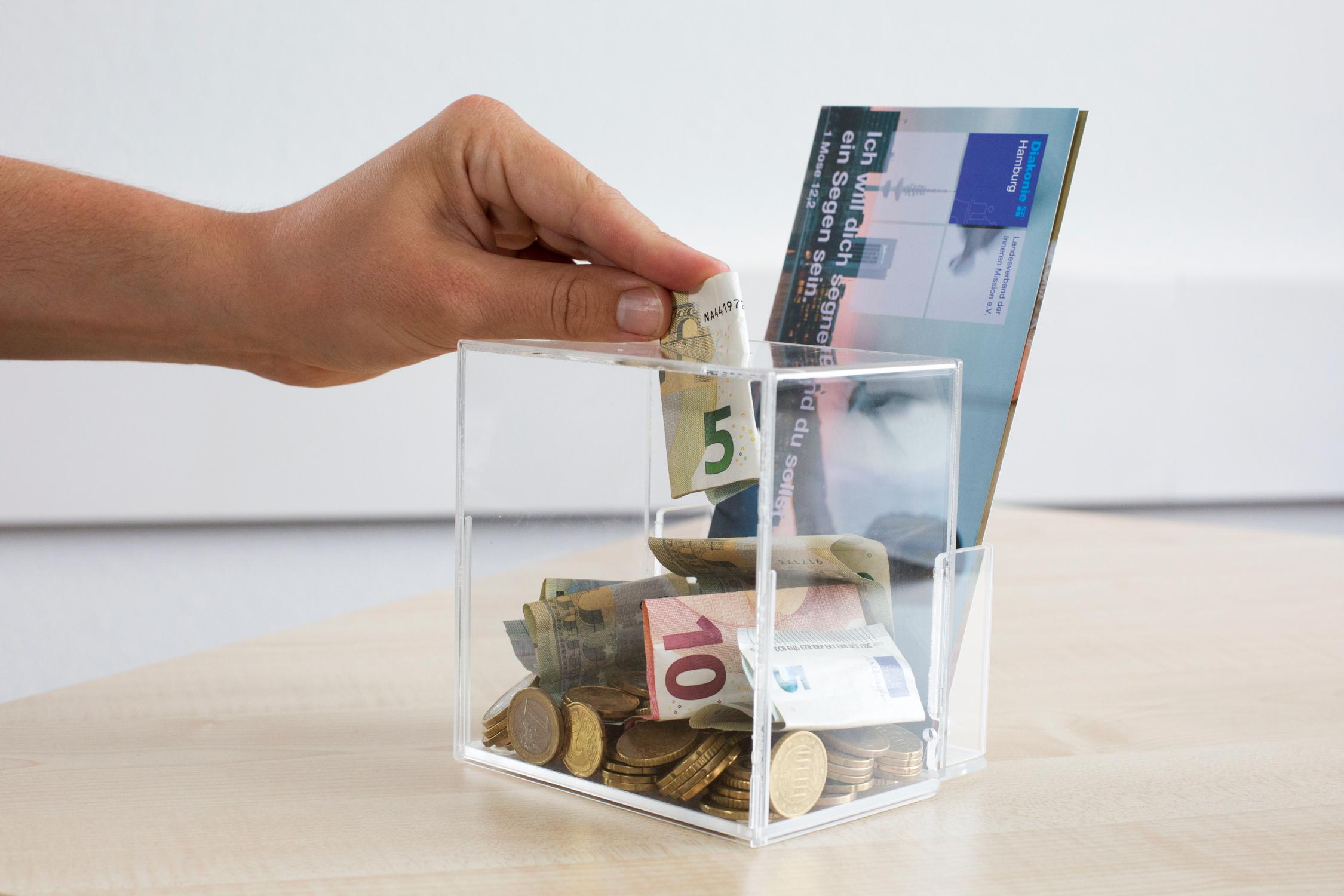 Eine Hand wirft einen Fünf-Euro-Schein in eine durchsichtige Spardose, in der bereits Geldscheine und Münzen liegen.