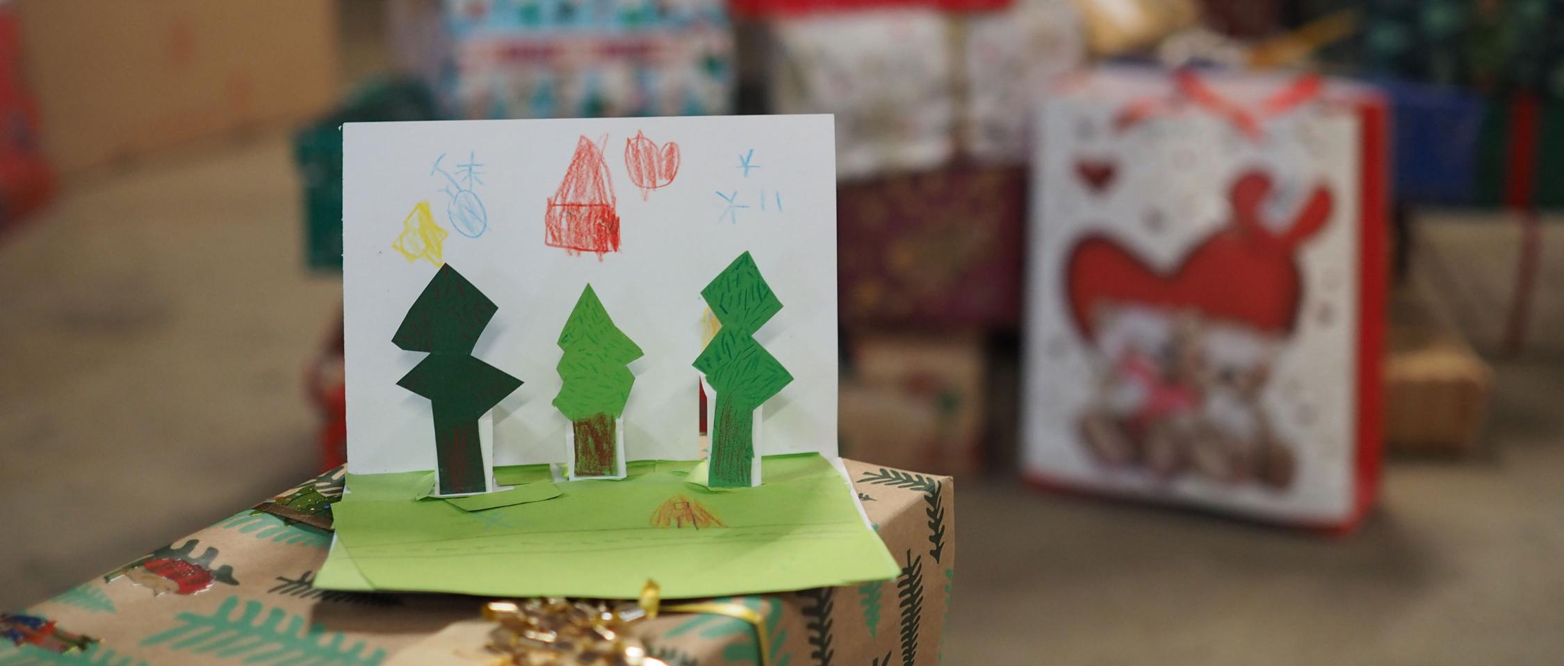 Eine selbst gebastelte Weihnachtskarte mit Tannenbäumen zum ausklappen steht auf einem Geschenk, im Hintergrund sind weitere Geschenke gestapelt zu sehen.