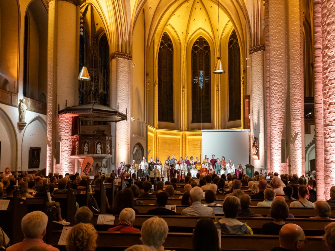 Innenraum einer Kirche mit Publikum auf Holzbänken und einer Gruppe von Menschen auf einer Bühne vor einem großen Fenster mit gotischen Spitzbögen