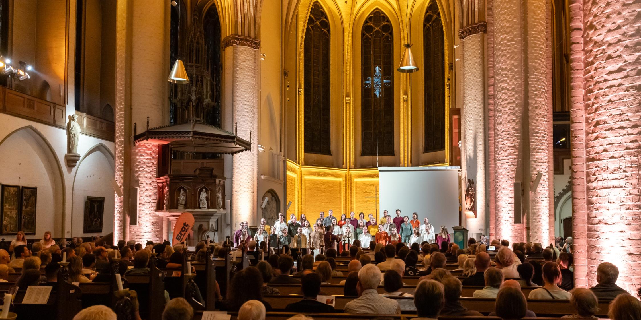 Innenraum einer Kirche mit Publikum auf Holzbänken und einer Gruppe von Menschen auf einer Bühne vor einem großen Fenster mit gotischen Spitzbögen