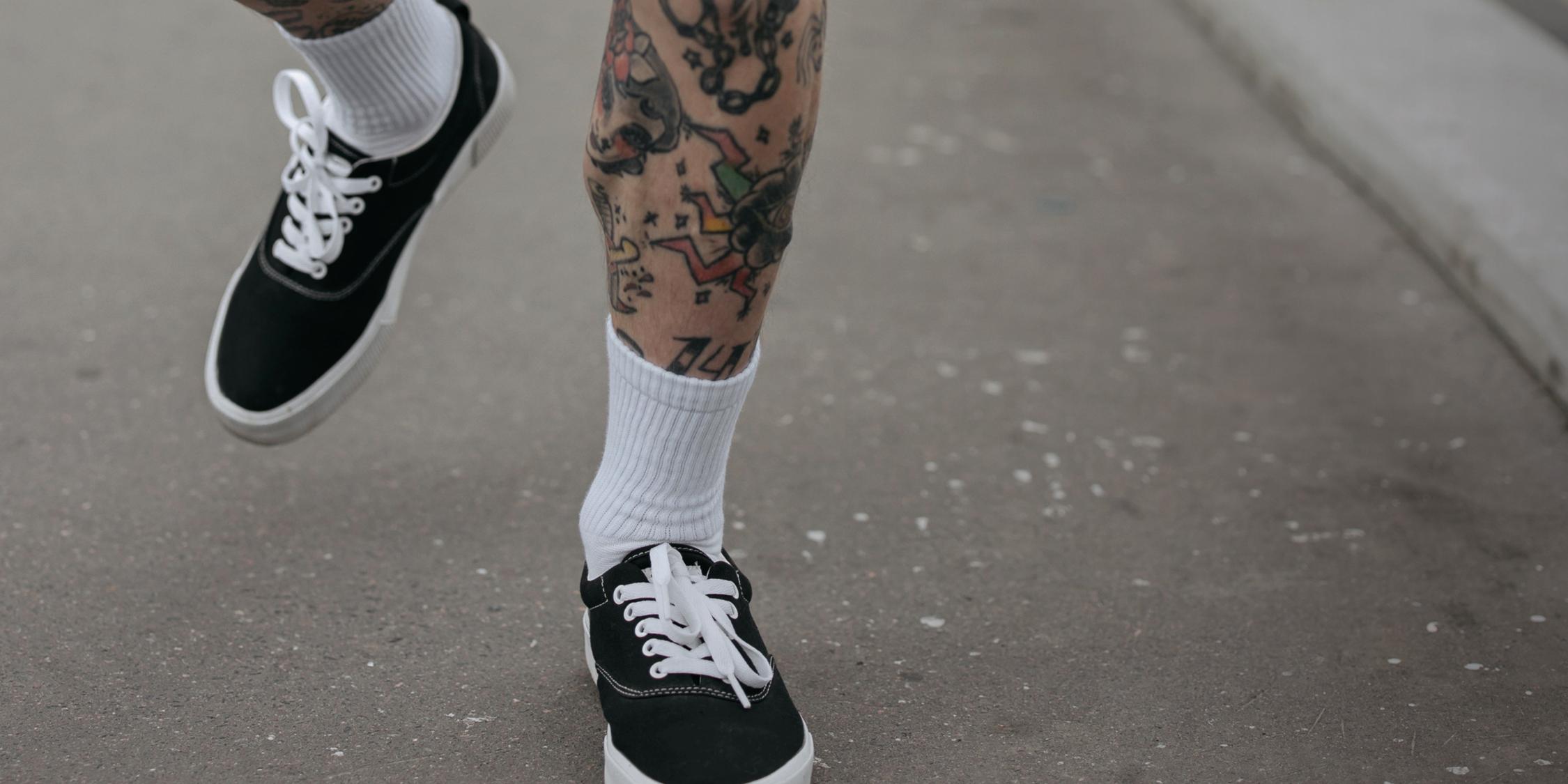 Tättoowierte Beine in schwarzen Sneakern und weißen Socken, die über eine Straße laufen.