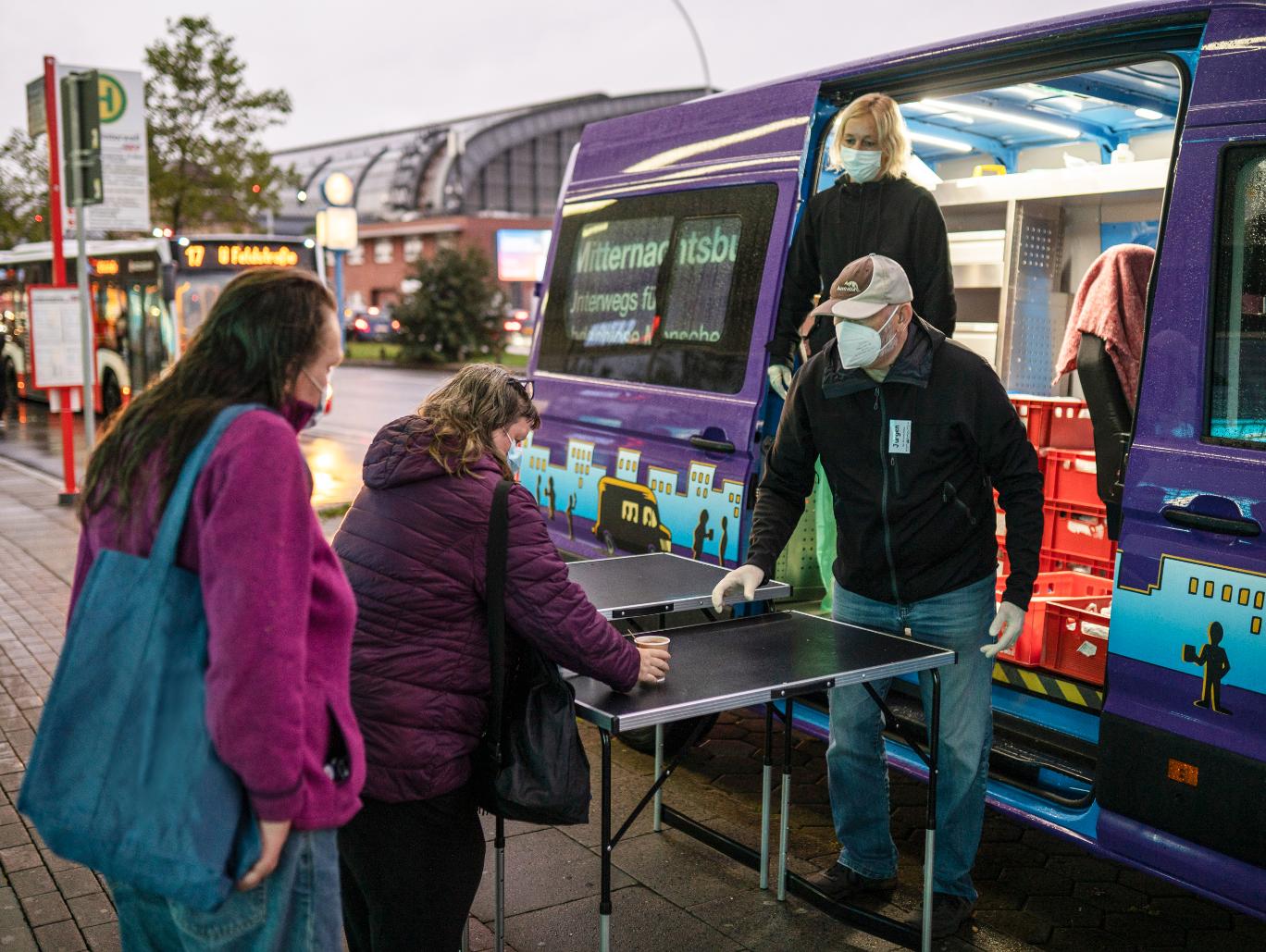 Eine Frau und ein Mann stehen am Mitternachtsbus und reichen zwei Frauen Kaffeebecher.