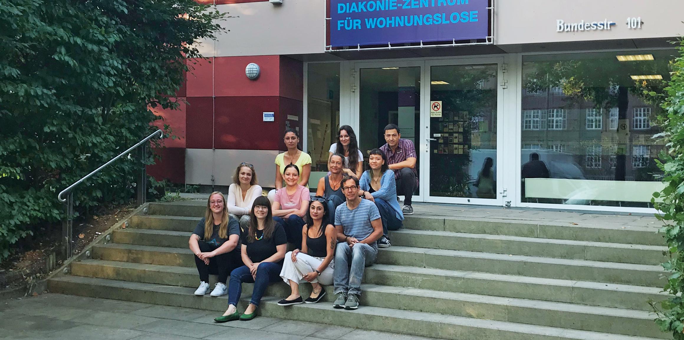Vor dem Diakonie-Zentrum für Wohnungslose sitzt eine diverse Gruppe von Menschen auf den Treppen.