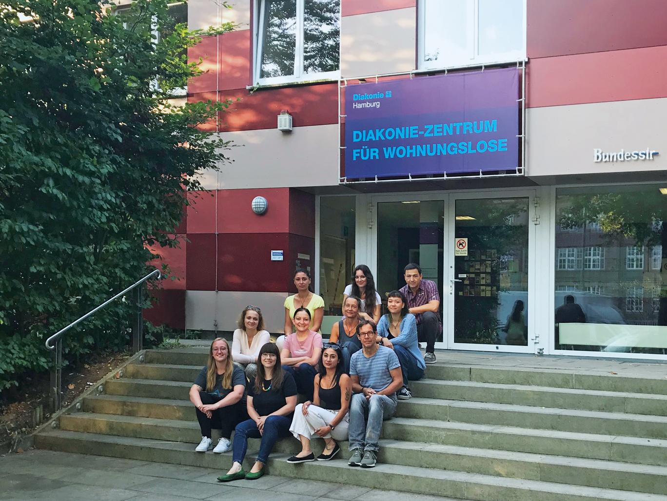 Vor dem Diakonie-Zentrum für Wohnungslose sitzt eine diverse Gruppe von Menschen auf den Treppen.