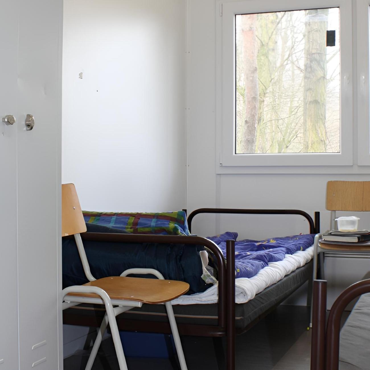 In einem kleinen Raum stehen zwei Betten, zwei Stühle und ein weißer Schrank.